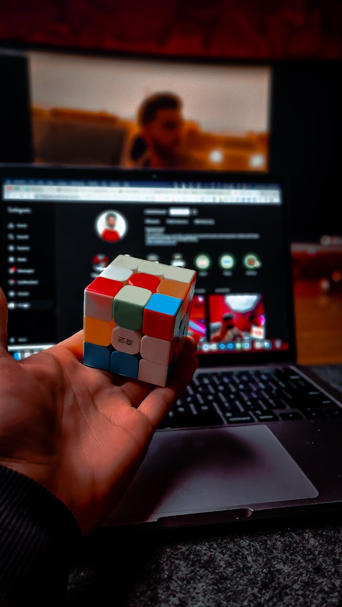 A hand holds a Rubiks Cube in front of a laptop with a blurred screen, indoors.